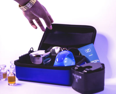 A blue Pitbull Deluxe Travel Case displayed open, showcasing its organized contents, including a silver shaver, a blue grooming tool, and a blue aftershave bottle. Nearby, a black Pitbull Platinum rinse stand and small glass bottles with liquid are visible. A hand wearing a gold watch reaches toward the case, highlighting its accessibility and premium design.
